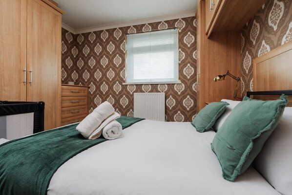 A neatly made bed with green pillows and a rolled white towel, wooden wardrobe, and a window with blinds.