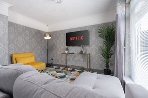 Ein modernes Wohnzimmer mit grauem Sofa, einem gelben Sessel, einem Couchtisch, einer Stehlampe, einem an der Wand montierten Fernseher und einem großen Fenster mit Jalousien.