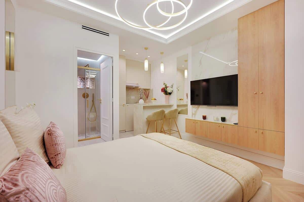 A modern bedroom with a large bed, a TV, a wooden wardrobe, and a small kitchenette.