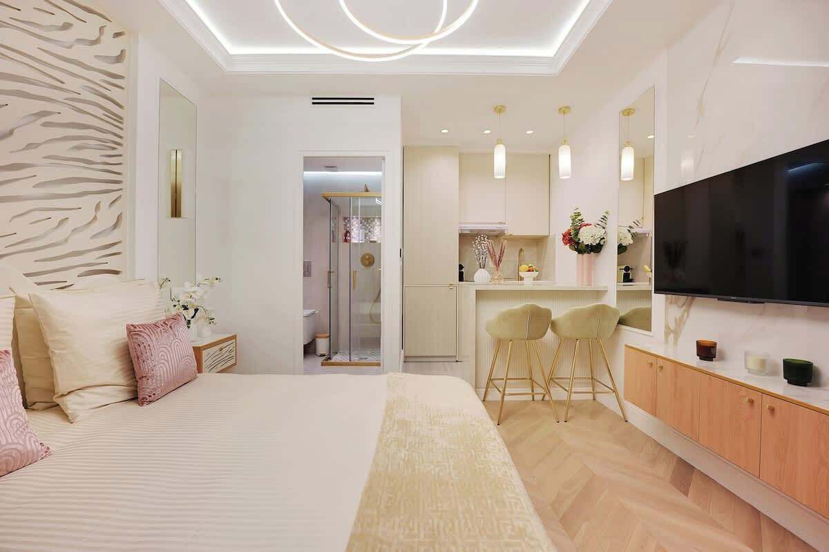 A modern hotel room with a large bed, a sitting area with a TV, and a kitchenette.