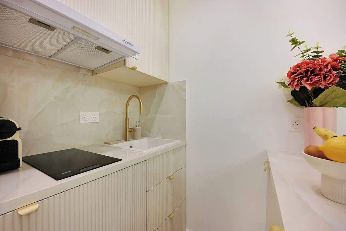 Een moderne keuken met een wit aanrecht, een zwart inductiefornuis, een gouden kraan en een witte spoelbak. Op het aanrecht staat een vaas met bloemen en een schaal met fruit.
