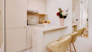 Eine moderne Küche mit weißer Arbeitsplatte, einem Spülbecken und einem Blumenstrauß. Zwei Stühle mit goldenen Beinen und eine weiße Wand mit Spiegel.