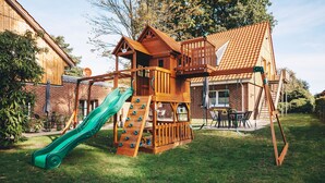 Een houten speelconstructie met een glijbaan en klimmuur, in een achtertuin met een stenen huis op de achtergrond.
