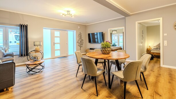 Een ruime woonkamer met een houten eettafel, zithoeken, een flatscreen televisie en een groot raam.