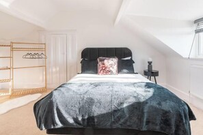 A bedroom with a bed, a black headboard, a white door, a wooden shelf unit, and a small table with a lamp. - 4 Bedroom Home w/ Free Parking - Tŷ Hapus Newport (Beechwood)