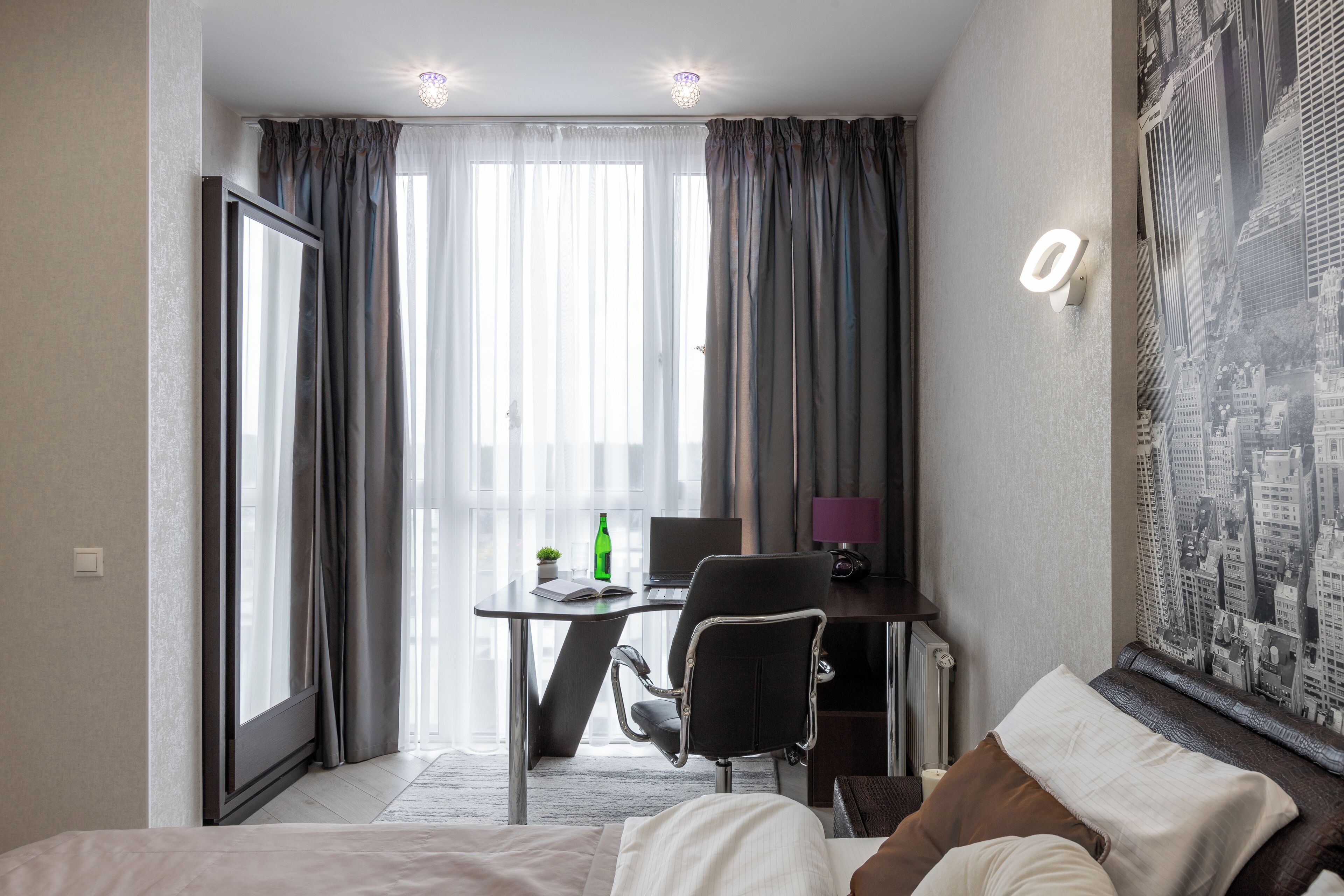 A hotel room with a bed, a desk with a laptop, a chair, and a window with curtains.