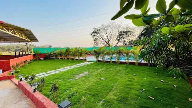 A well-maintained lawn with a paved walkway, surrounded by greenery and a building with a red exterior. - Amantran Farms and Resort (Karjat)