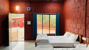 A hotel room with a bed, a window with curtains, a view of the outside, and a brick wall. - Amantran Farms and Resort (Karjat)