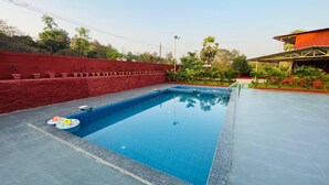 A rectangular swimming pool with a clear blue water surface, surrounded by a red wall and a paved deck. - Amantran Farms and Resort (Karjat)
