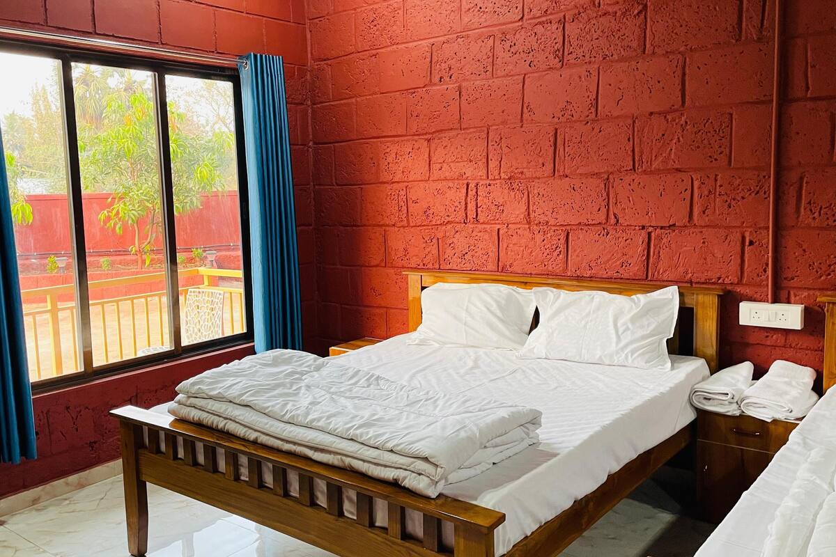 A bedroom with a wooden bed, white bedding, a window with blue curtains, and a red brick wall.