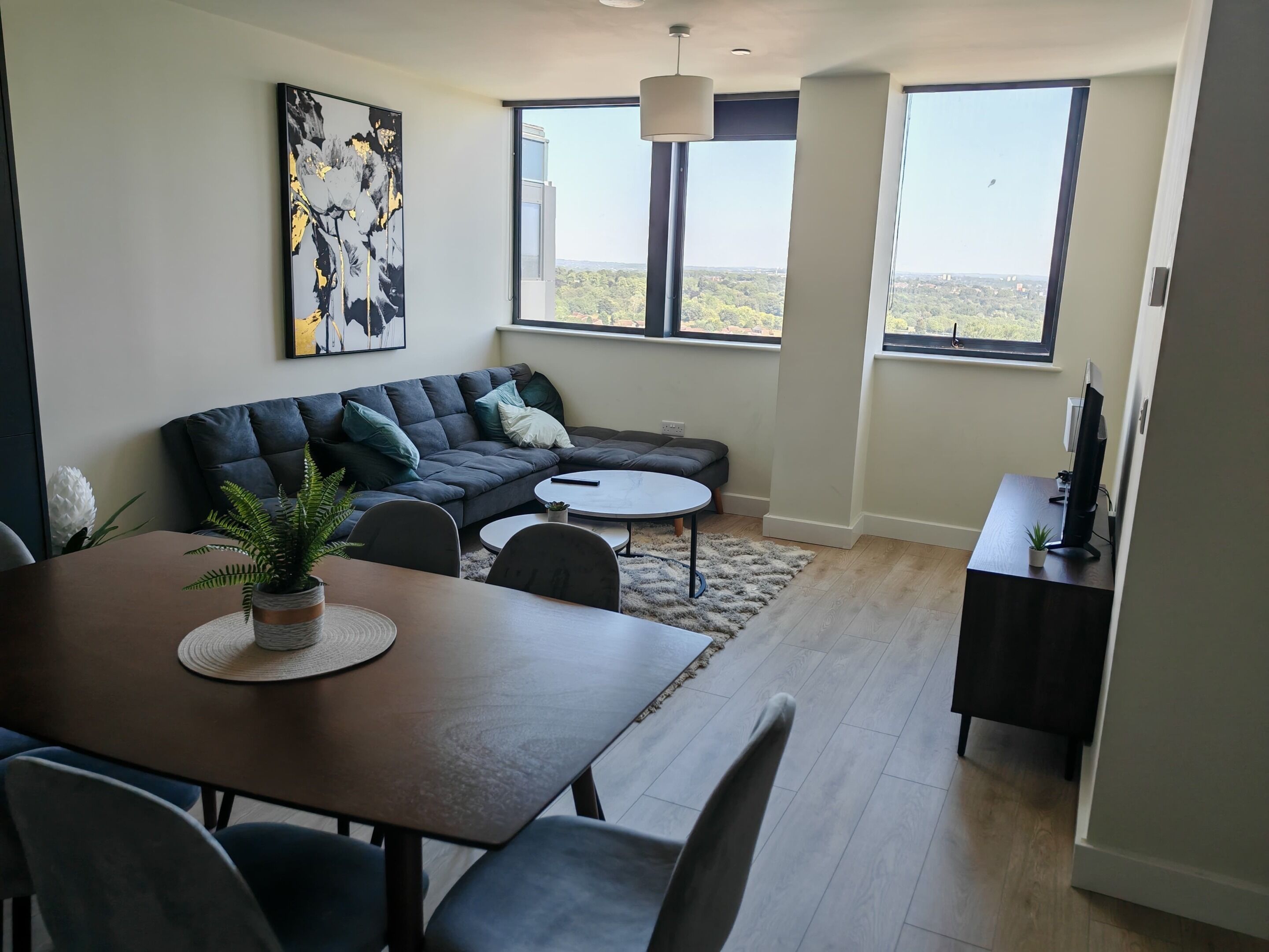 A modern living room with a dark gray sectional sofa, a round coffee table, and a dining table with chairs. There is a large window offering a view of the landscape.