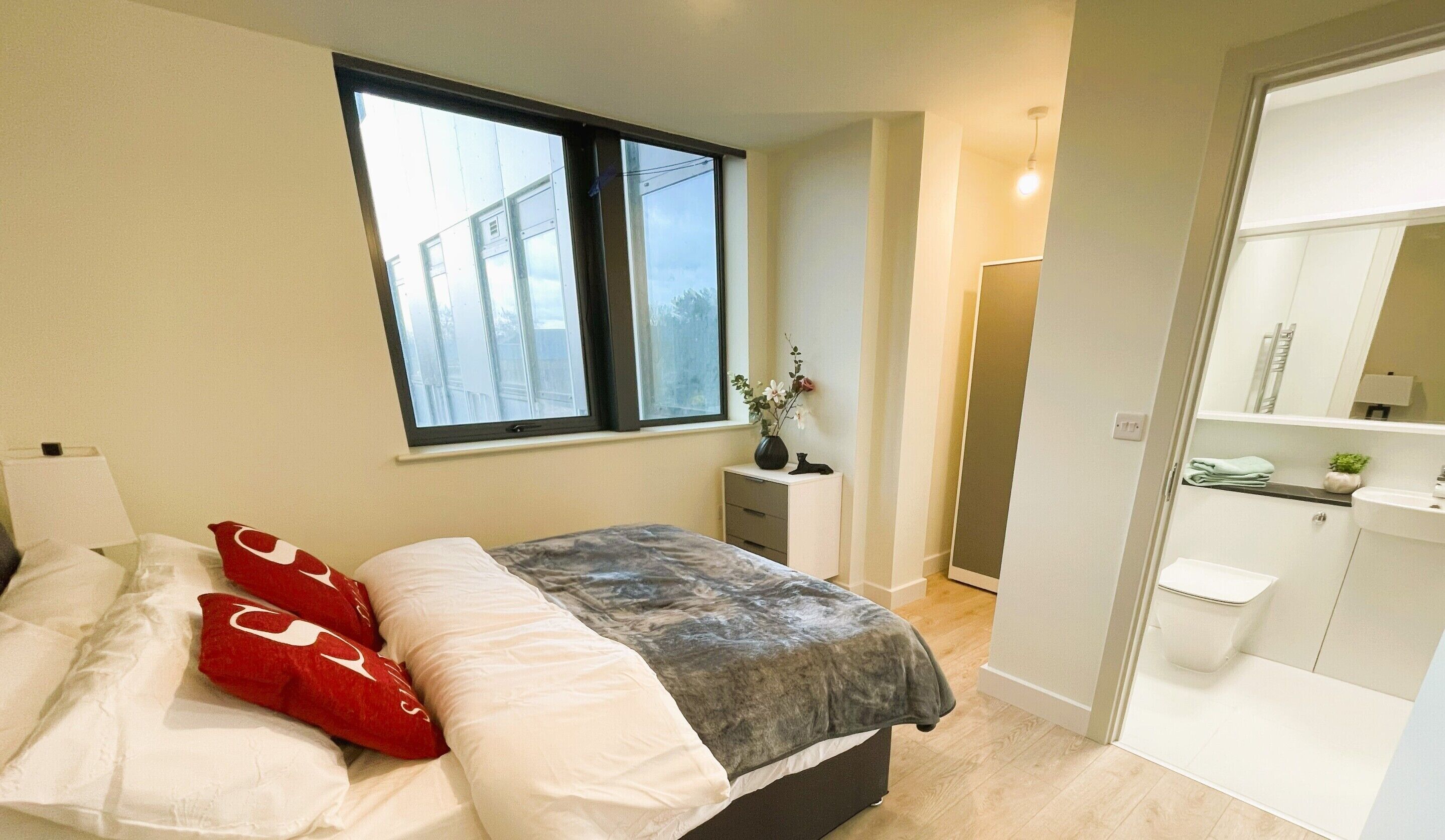A bedroom with a bed, a window, a bedside table, and a bathroom with a mirror and sink.