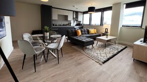Un salon moderne comprenant un canapé d’angle, une table à manger et des chaises. On y trouve également un coin cuisine avec des placards et une fenêtre.