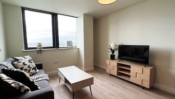 Ein modernes Wohnzimmer mit einer Couch, einem Couchtisch, einem Fernseher auf einem Holzboden und einem Fenster mit Blick auf die Stadt.