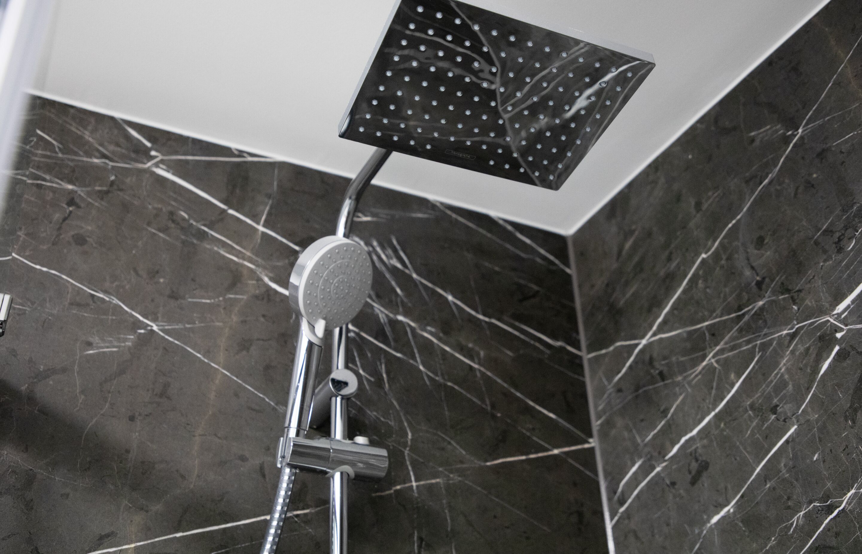 Una ducha moderna con cabezal cuadrado, accesorios de cromo y pared de mármol con vetas blancas.