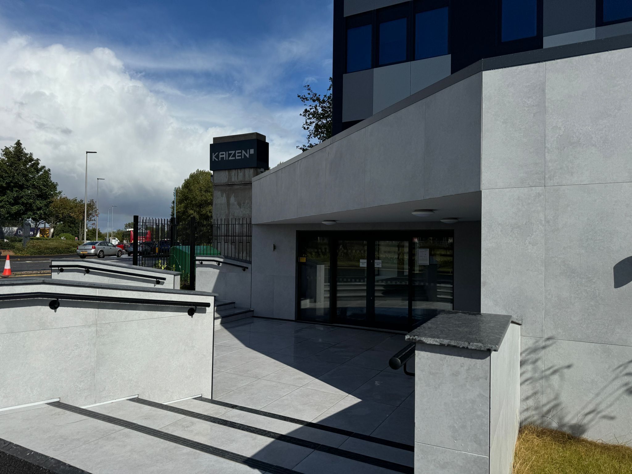 Een modern gebouw met een 'KAIZEN' bord, een overdekte ingang en een verhard gebied met treden naar de ingang.