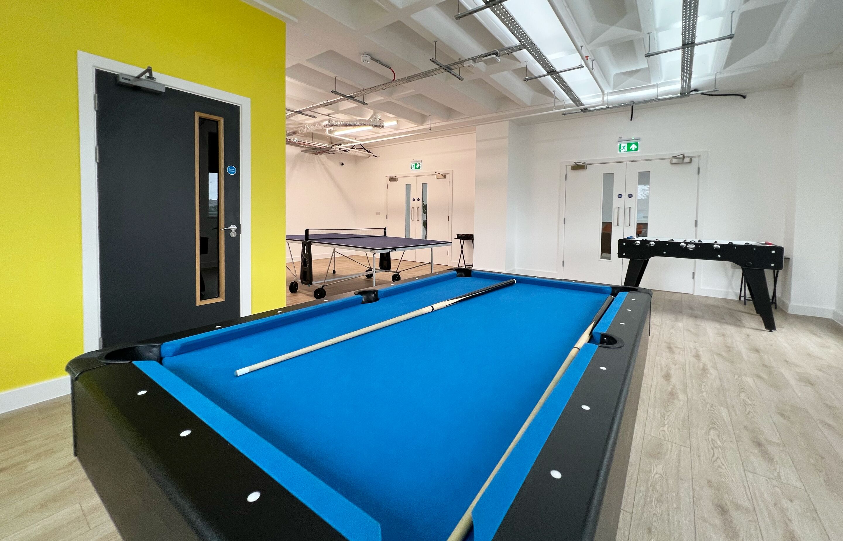 A pool table with a blue felt surface, a ping pong table, and a foosball table in a room with yellow and white walls and wooden flooring.