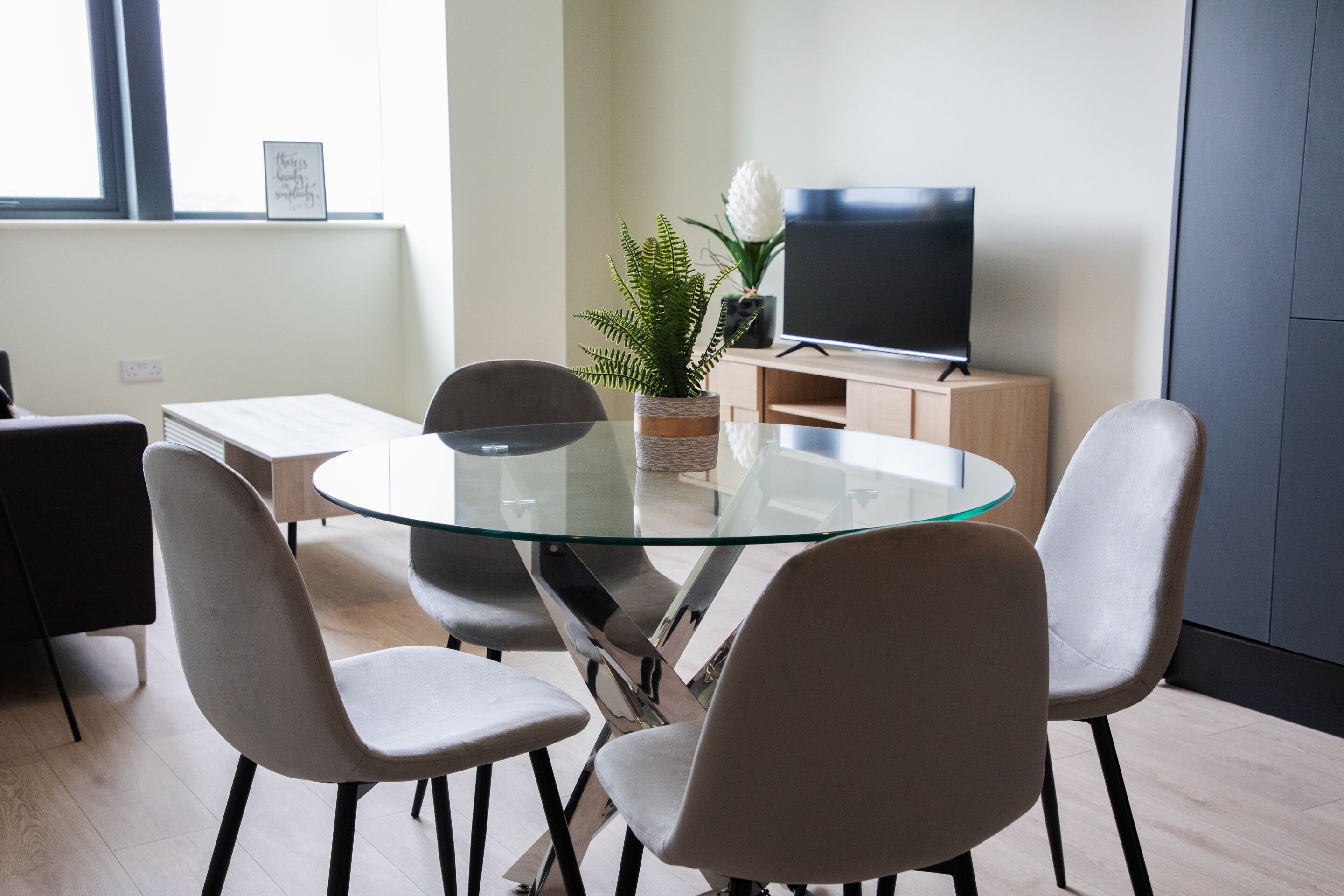 A modern dining area with a glass table, four chairs, a television on a wooden cabinet, and a potted plant.