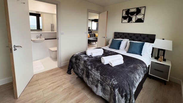 Une chambre moderne avec un grand lit, des tables de chevet, une petite table d’appoint, un luminaire suspendu et une salle de bain visible à travers une porte ouverte.