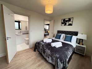 A modern bedroom with a large bed, bedside tables, a nightstand, a hanging light fixture, and a bathroom visible through an open door. - Tha Kaizen (West Bromwich)