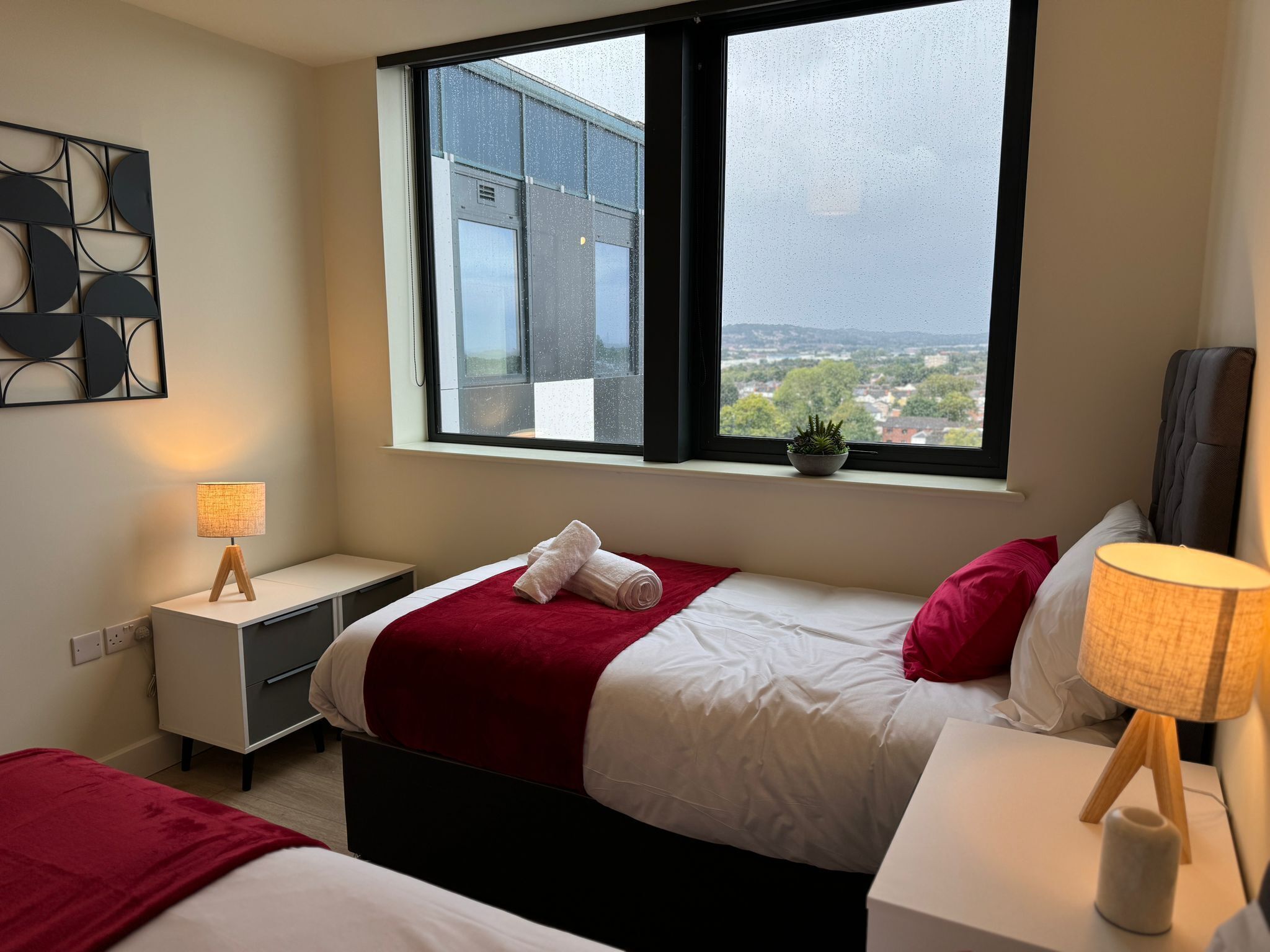 Une chambre d’hôtel moderne avec une grande fenêtre, un lit recouvert d’une couverture rouge, une table de chevet avec une lampe et une petite plante en pot posée sur le rebord de la fenêtre.
