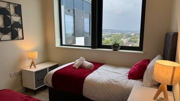 Une chambre d’hôtel moderne avec une grande fenêtre, un lit recouvert d’une couverture rouge, une table de chevet avec une lampe et une petite plante en pot posée sur le rebord de la fenêtre.