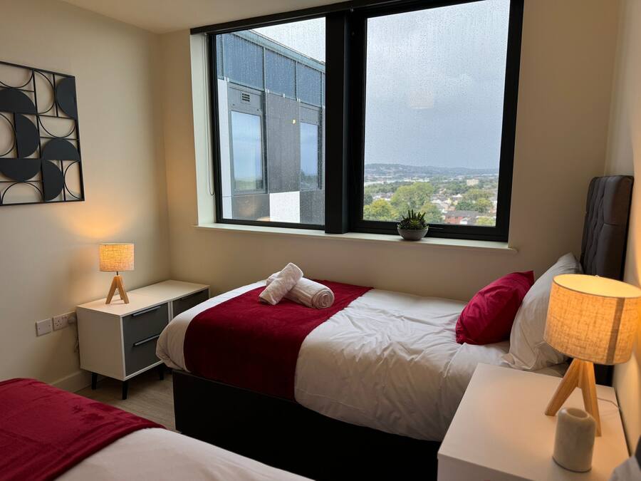 Une chambre d’hôtel moderne avec une grande fenêtre, un lit recouvert d’une couverture rouge, une table de chevet avec une lampe et une petite plante en pot posée sur le rebord de la fenêtre.