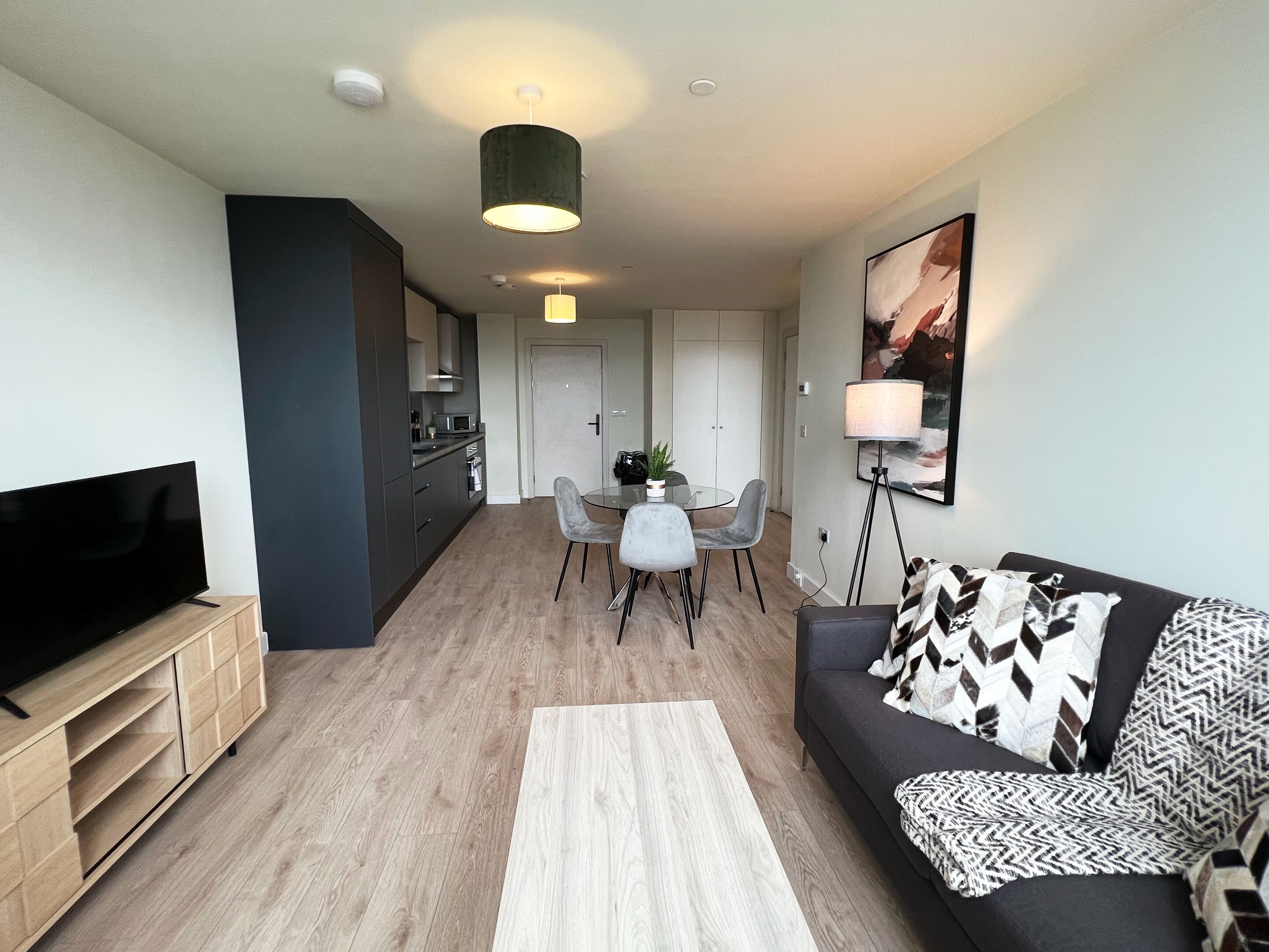 Eine moderne Wohnung mit Flachbildfernseher, einer Essecke mit Glastisch und Stühlen, einem Sofa mit gemusterten Kissen und einer Küche mit dunklen Schränken.