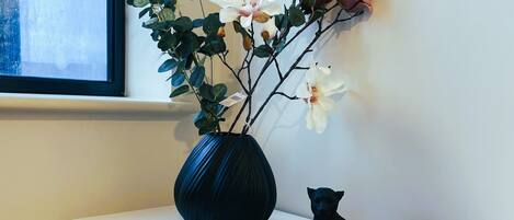 Un vase rempli de fleurs et une statuette de chat noir sont posés sur une table blanche, devant un mur.