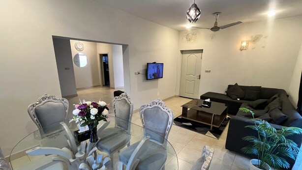 Ein Wohnzimmer mit einem Glastisch, grau gepolsterten Sesseln, einem schwarzen Sofa, einem Deckenventilator, einem Fernseher und einer Topfpflanze.