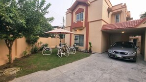 Ein zweistöckiges Haus mit einem Auto auf der Einfahrt, einer Terrasse mit Stühlen und einem Sonnenschirm sowie einem kleinen Garten.