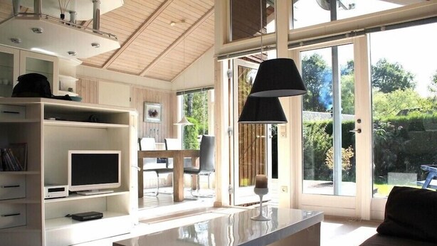 A modern living room with a TV, bookshelves, a dining area, and a large sliding glass door to the outdoors. - FeWoHuett - cozy vacation home in the Hüttener mountains near the Baltic Sea (Brekendorf)