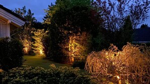 A garden with illuminated trees and shrubs, a paved path, and a building in the background. - FeWoHuett - cozy vacation home in the Hüttener mountains near the Baltic Sea (Brekendorf)