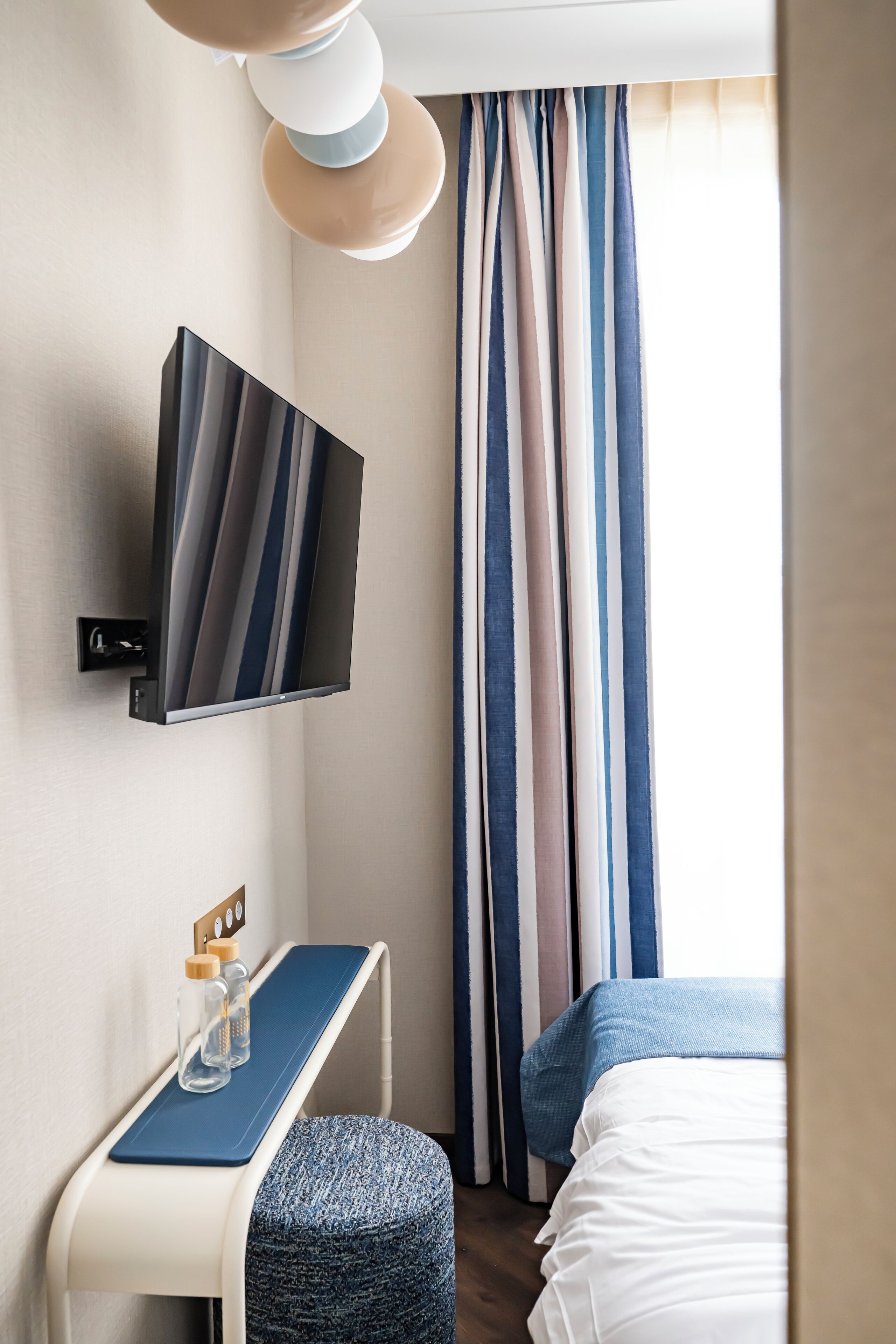 Una habitación de hotel moderna con una televisión de pantalla plana montada en la pared, un escritorio pequeño con una silla, y una cama con cabecera azul.