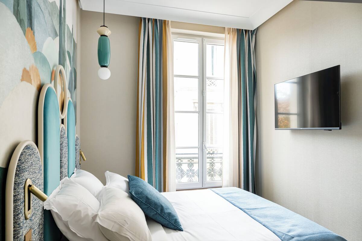 Une chambre à coucher avec un lit, un téléviseur fixé au mur, une fenêtre avec des rideaux et un mur orné d’une fresque décorative.