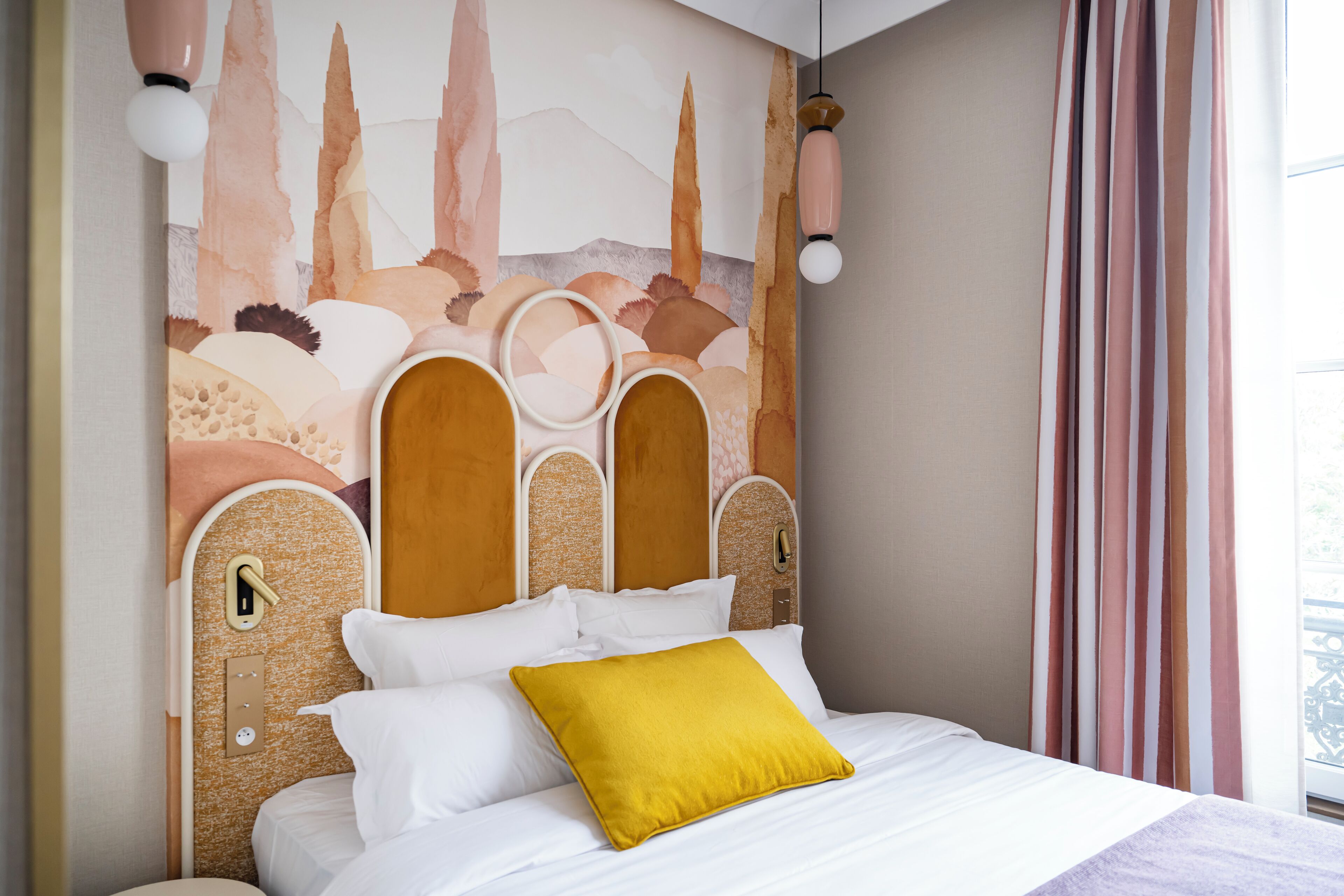 Un lit avec une literie blanche et un oreiller jaune moutarde, une tête de lit au motif texturé et un mur orné d’un paysage peint.