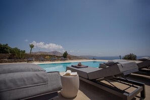 A poolside area with lounge chairs, a small table with a hat and books, and a clear view of the sea and mountains in the background.