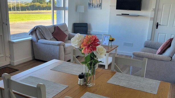 Un salon moderne avec une table à manger en bois, des canapés gris recouverts de tissu et un téléviseur à écran plat fixé au mur.