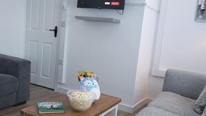 Un salon avec un téléviseur à écran plat fixé au mur, une table basse en bois, deux canapés gris et une petite table sur laquelle se trouvent un bol de pop-corn et un livre.