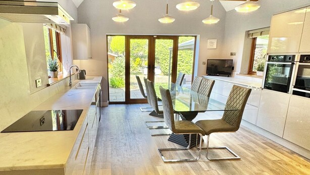A modern kitchen with a dining area, featuring a large island, pendant lighting, and a view through glass doors.