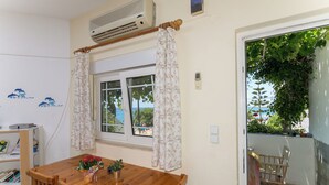 A room with a wooden dining table, chairs, a window with curtains, an air conditioning unit, and a view of greenery outside. - Crete Essence Villas-Makrygialos (Ierapetra)