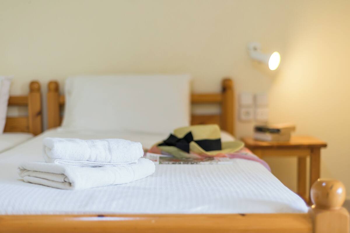 Uma cama bem arrumada com toalhas dobradas, um chapéu colorido e uma mesa de cabeceira com abajur.