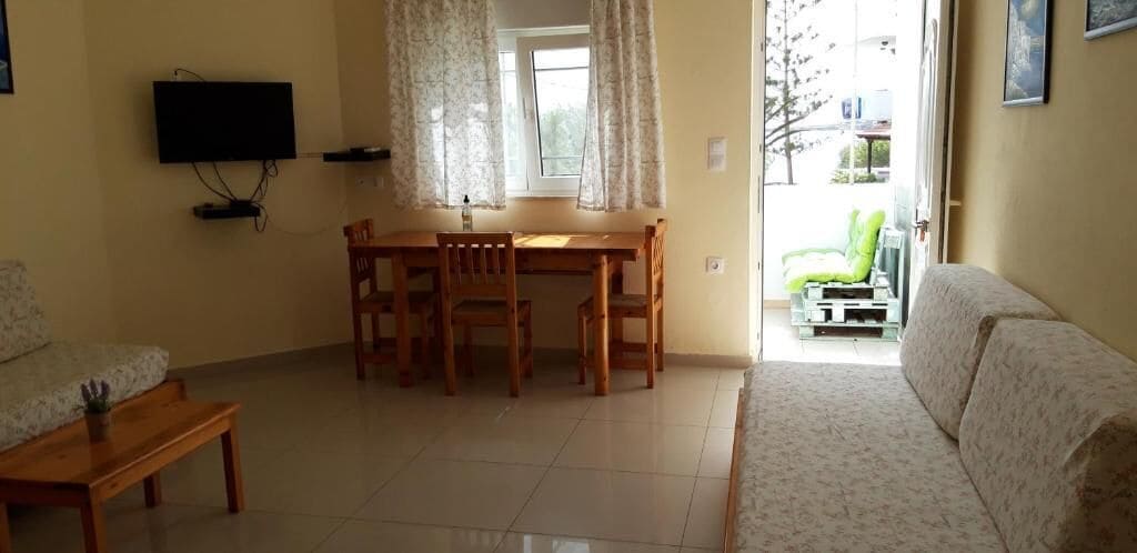 A living room with a wooden dining table, two chairs, a sofa, and a television mounted on the wall.