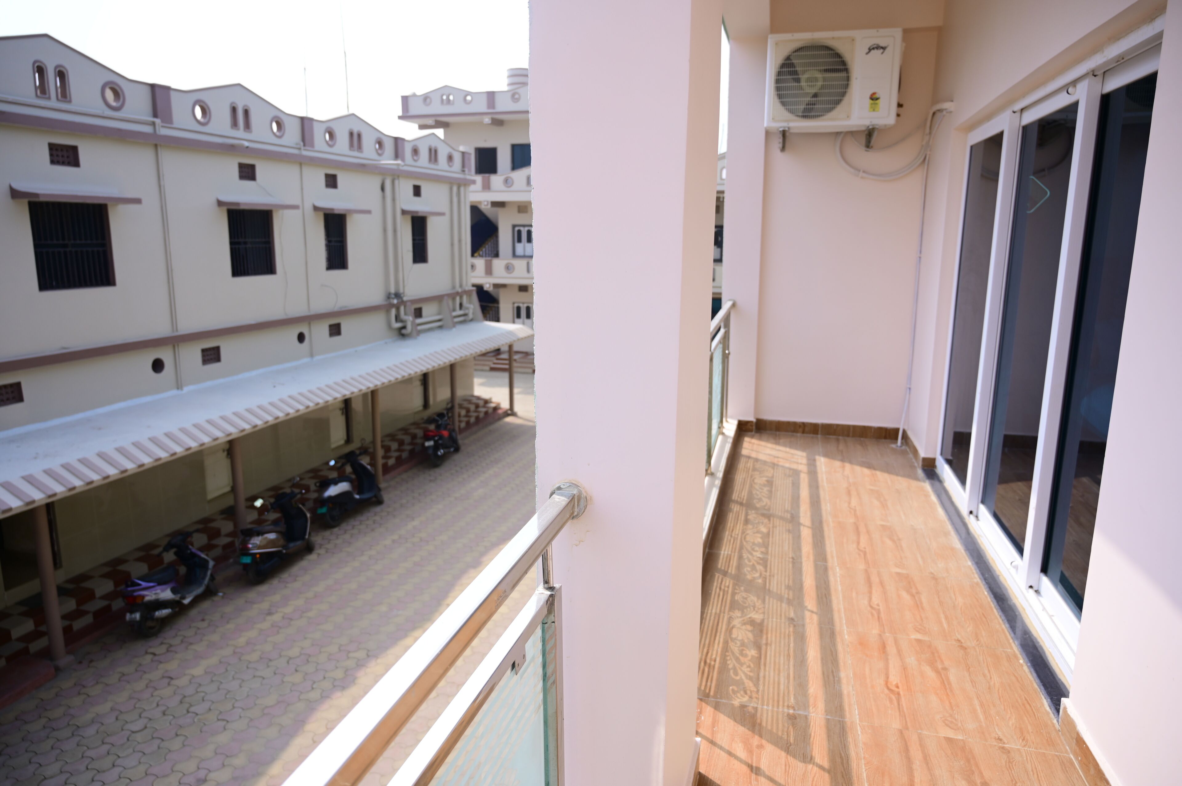 Ein Balkon mit Fliesenboden, ein Fenster mit Blick auf abgestellte Roller und eine Klimaanlage an der Wand.