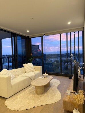 A modern living room with a large sofa, a coffee table, and a view of a cityscape through floor-to-ceiling windows.