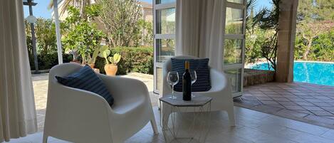 Uma área de estar moderna ao ar livre, com cadeiras brancas e mesa de vidro, com vista para uma piscina e vegetação exuberante.