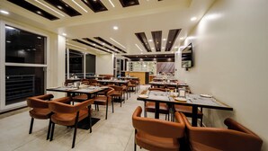 A modern dining area with multiple tables and chairs, recessed ceiling lighting, and a large window on the left. - Abhilashi Inn Kasol (Kasol)