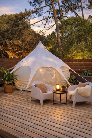 A tent with a white canopy, two white chairs, a small table with a lantern, and potted plants on a wooden deck.