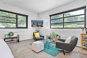 Een moderne woonkamer met grote ramen, een grijze bank, een witte poef en een salontafel.