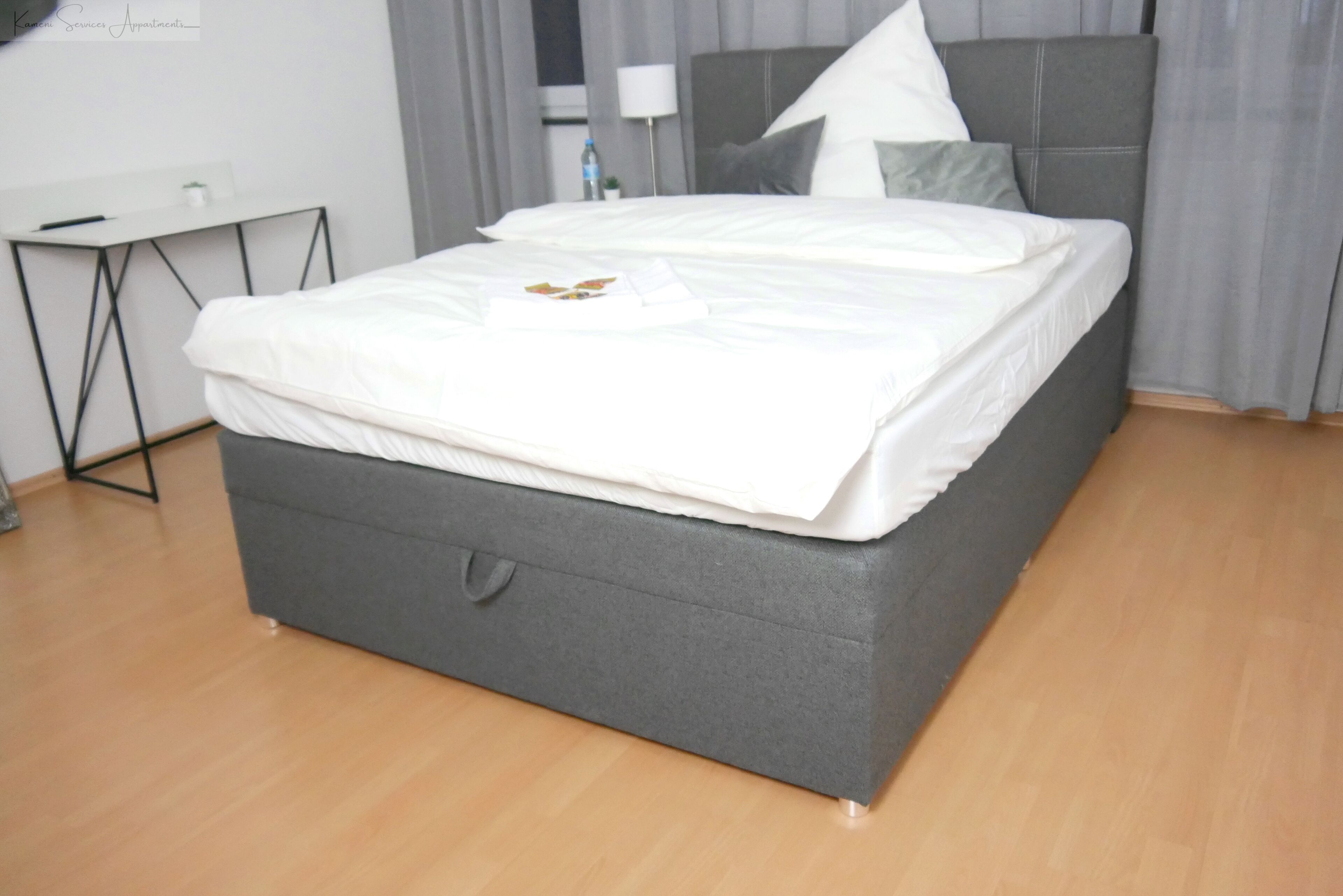 Ein ordentlich bezogenes Bett mit grauem Kopfteil und Fußteil, ein kleiner Nachttisch mit Lampe und eine weiße Wand mit Steckdose.